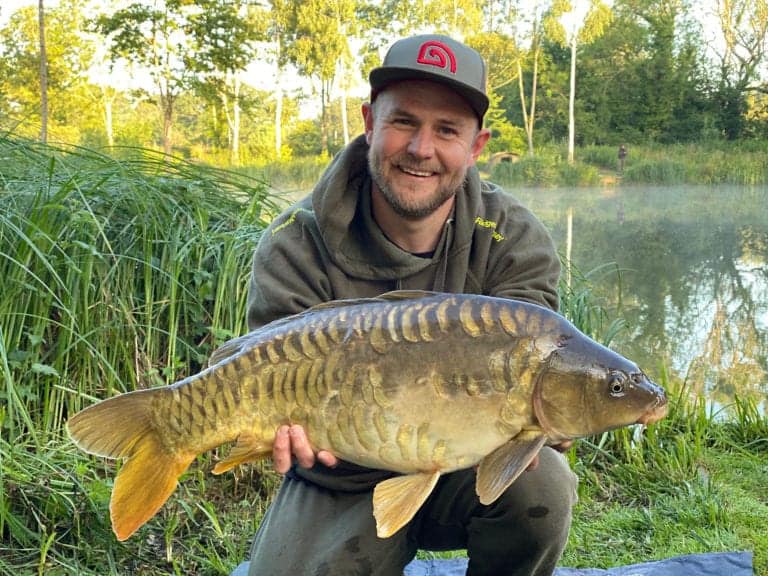 Smiling angler holding large mirror carp by lake.
